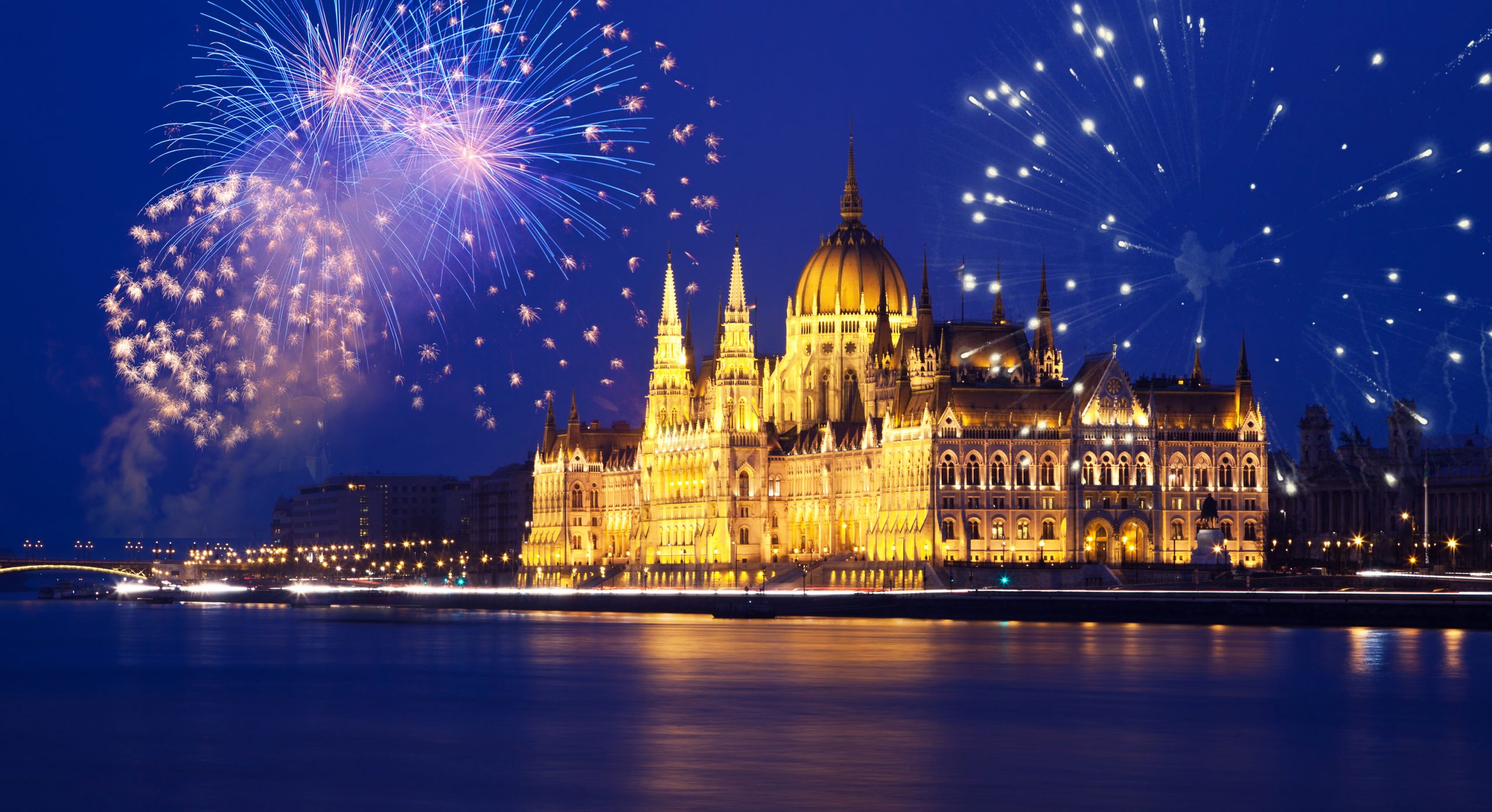 Espectaculares fuegos artificiales iluminando el cielo nocturno sobre el edificio del Parlamento de Budapest, reflejándose en el río Danubio durante la celebración de Fin de Año. Las luces del edificio y los destellos de los fuegos crean una atmósfera mágica y festiva.